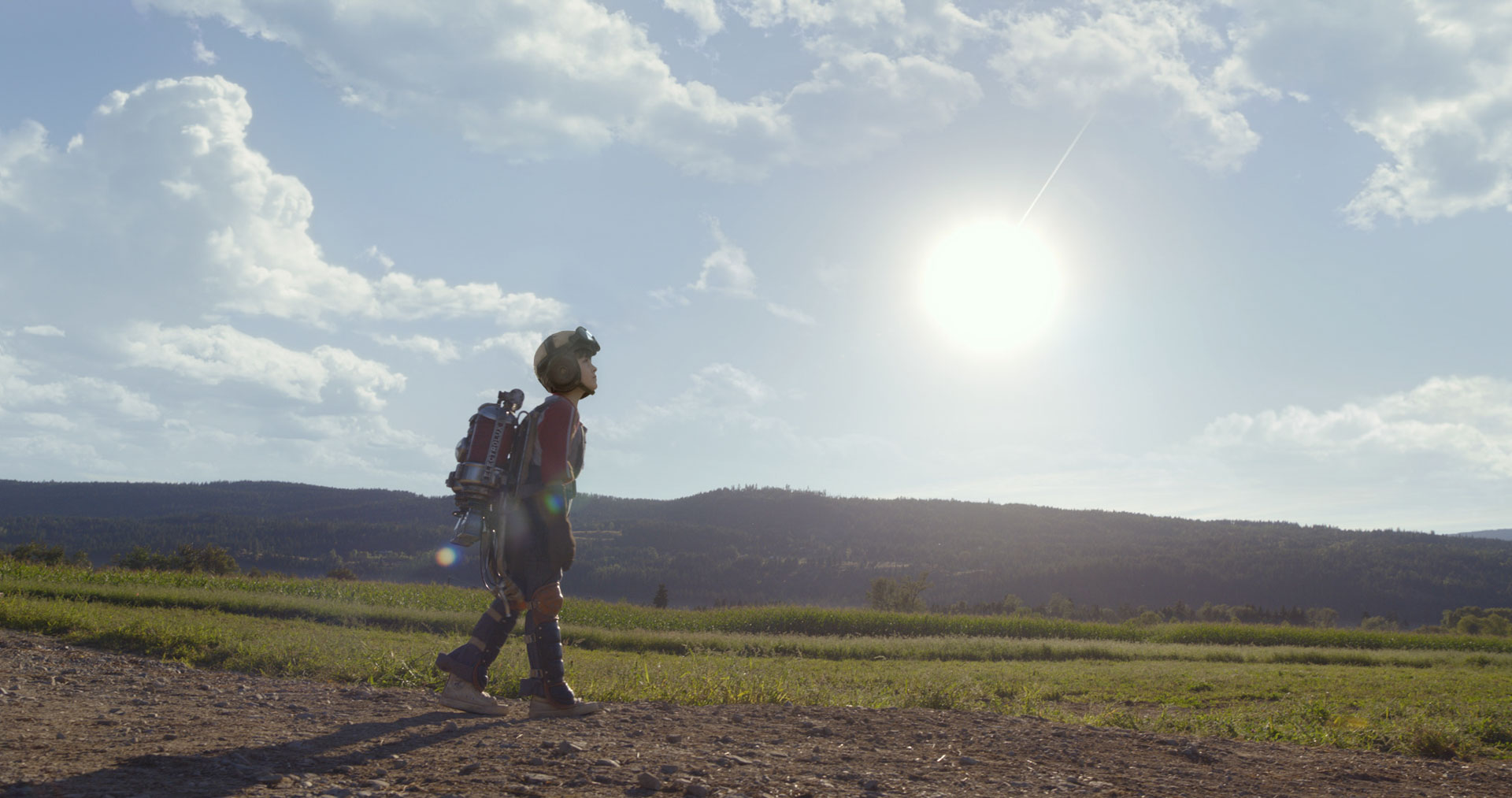 A child in makeshift armor and a helmet walks alone down a dirt path through a sunlit field, wearing a large homemade jetpack. The bright sun hangs low in the sky, casting long shadows as the child gazes toward the horizon with determination.