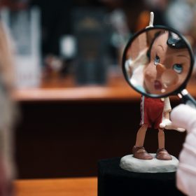 An archivist’s white-gloved hand is seen holding a magnifying glass up to a small animator’s model of Pinocchio (whose right arm is raised, pointing upward, and who is wearing red trousers and brown shoes). The character’s face appears enlarged through the magnifying glass.