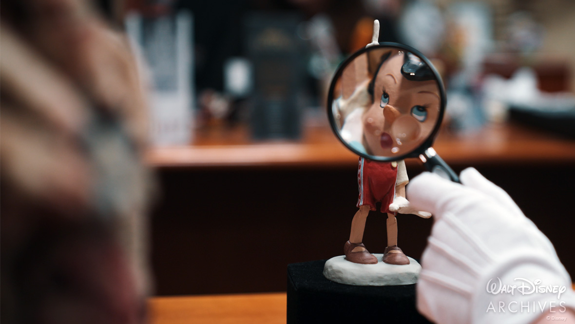 An archivist’s white-gloved hand is seen holding a magnifying glass up to a small animator’s model of Pinocchio (whose right arm is raised, pointing upward, and who is wearing red trousers and brown shoes). The character’s face appears enlarged through the magnifying glass.