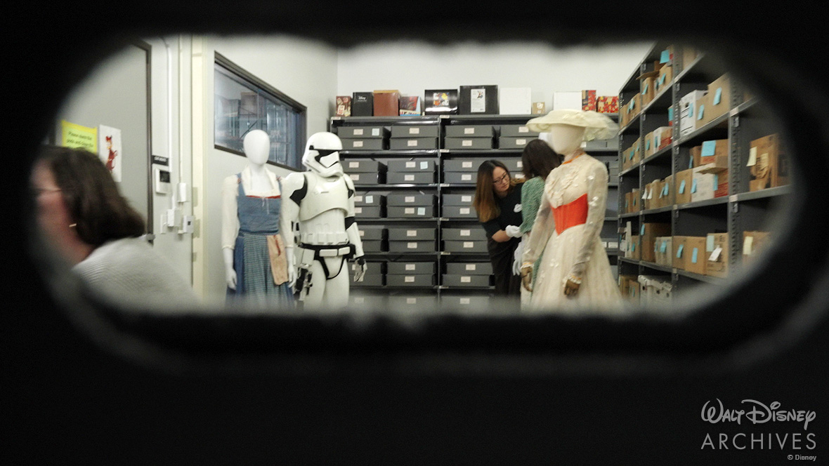 From the opening of a box, the perspective of the Pinocchio model looks out to view shelves of archival boxes, with a few costumes on display: Belle’s blue village dress from Beauty and the Beast (2017), a white Stormtrooper uniform from the Star Wars films, and Mary Poppins’ white “Jolly Holiday” dress and hat from the 1964 film. Two archives staff members are seen at work in the background. 
