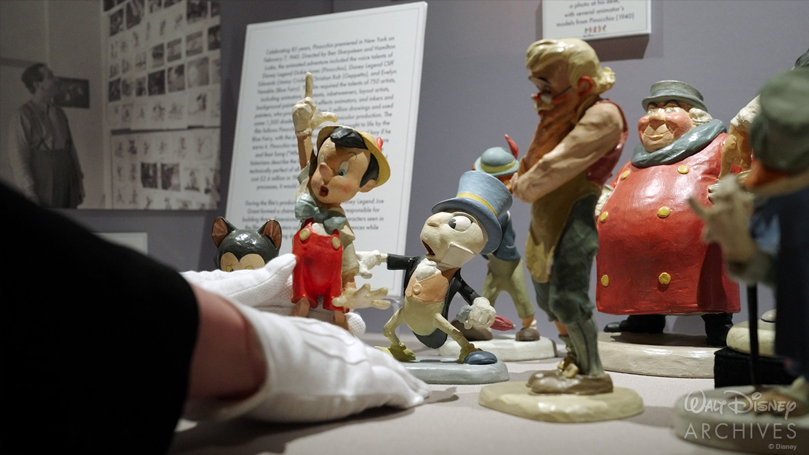 An archivist’s white-gloved hands are seen placing the Pinocchio model in a display case alongside similar models created for the production of the 1940 film, including the black-and-white cat Figaro; Jiminy Cricket, wearing a blue top hat and holding an umbrella; Geppetto, who is wearing glasses and has his right arm and hand to his chin; the Coachman, who is wearing a red coat, blue scarf, and green top hat; and J. Worthington Fowlfellow, the fox.