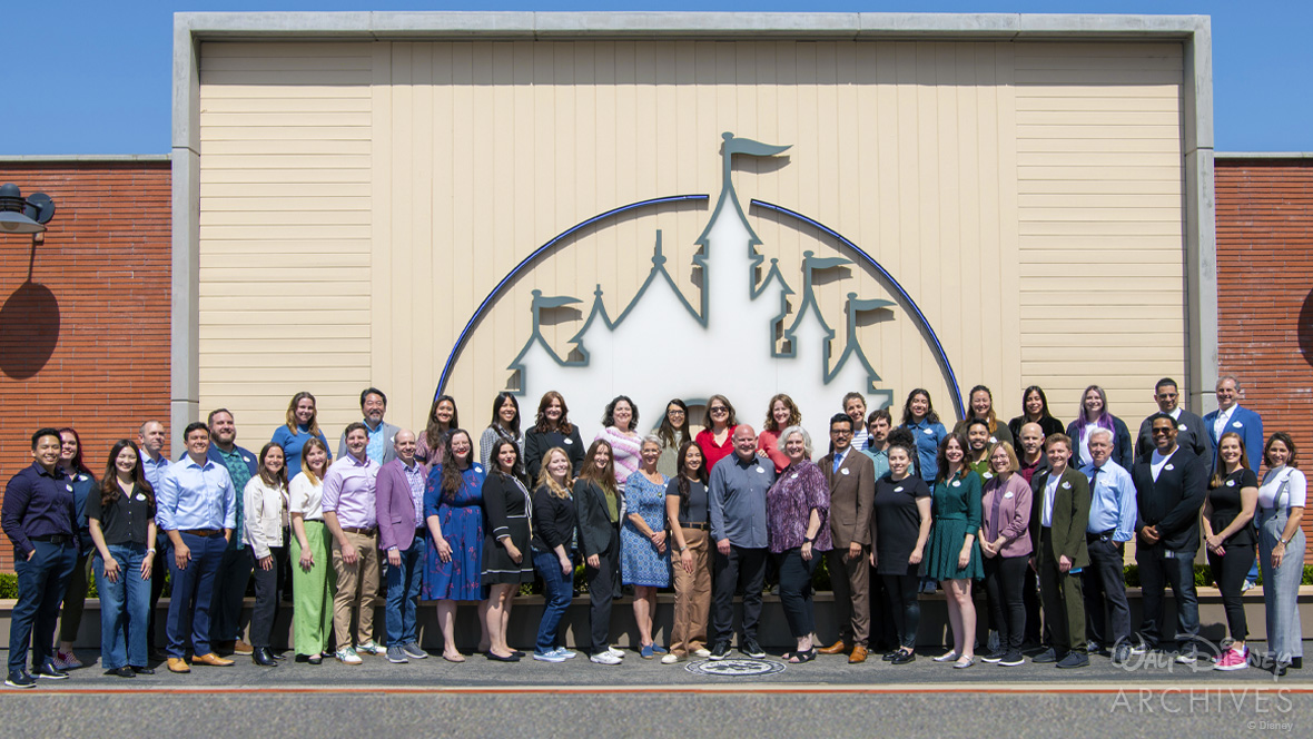 Outside on the backlot of The Walt Disney Studios, forty-six members of the Walt Disney Archives organization pose for a group photo, arranged in two rows, in front of a large mounted depiction of Sleeping Beauty Castle, which is set in white, flanked by two brick-faced edifices.