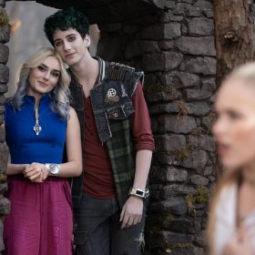 Still from Zombies 4: Dawn of the Vampires. Victor, wearing a red and black shirt, and Nova, wearing a red and white zip-up jacket, have a conversation in the foreground. Zed, wearing a green vest over a red shirt, and Addison, wearing a blue shirt and magenta pants, lean on one another as they look at Victor and Nova from the background.