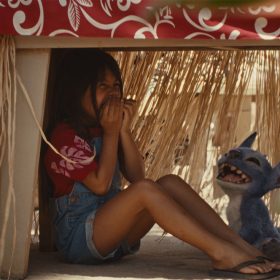 Lilo (Maia Kealoha) and Stitch from the live-action reimagining of Lilo & Stitch hiding under a table and laughing.