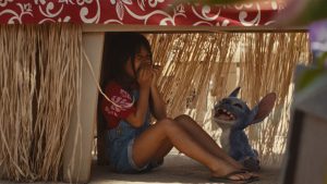 Lilo (Maia Kealoha) and Stitch from the live-action reimagining of Lilo & Stitch hiding under a table and laughing.