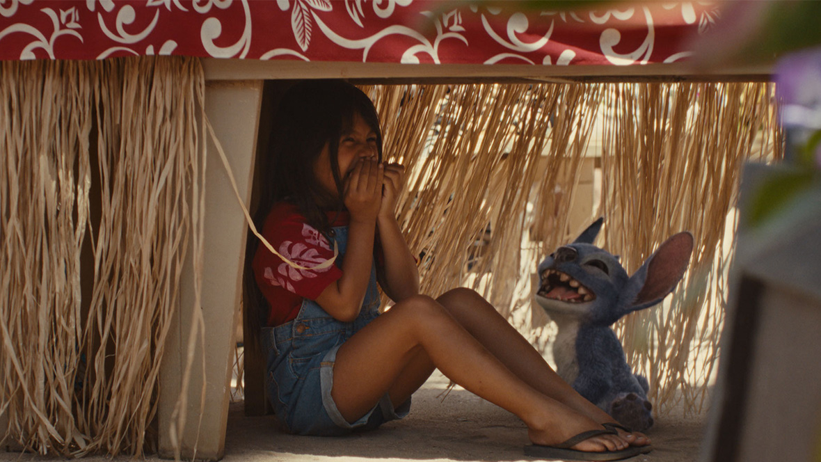 Lilo (Maia Kealoha) and Stitch from the live-action reimagining of Lilo & Stitch hiding under a table and laughing.