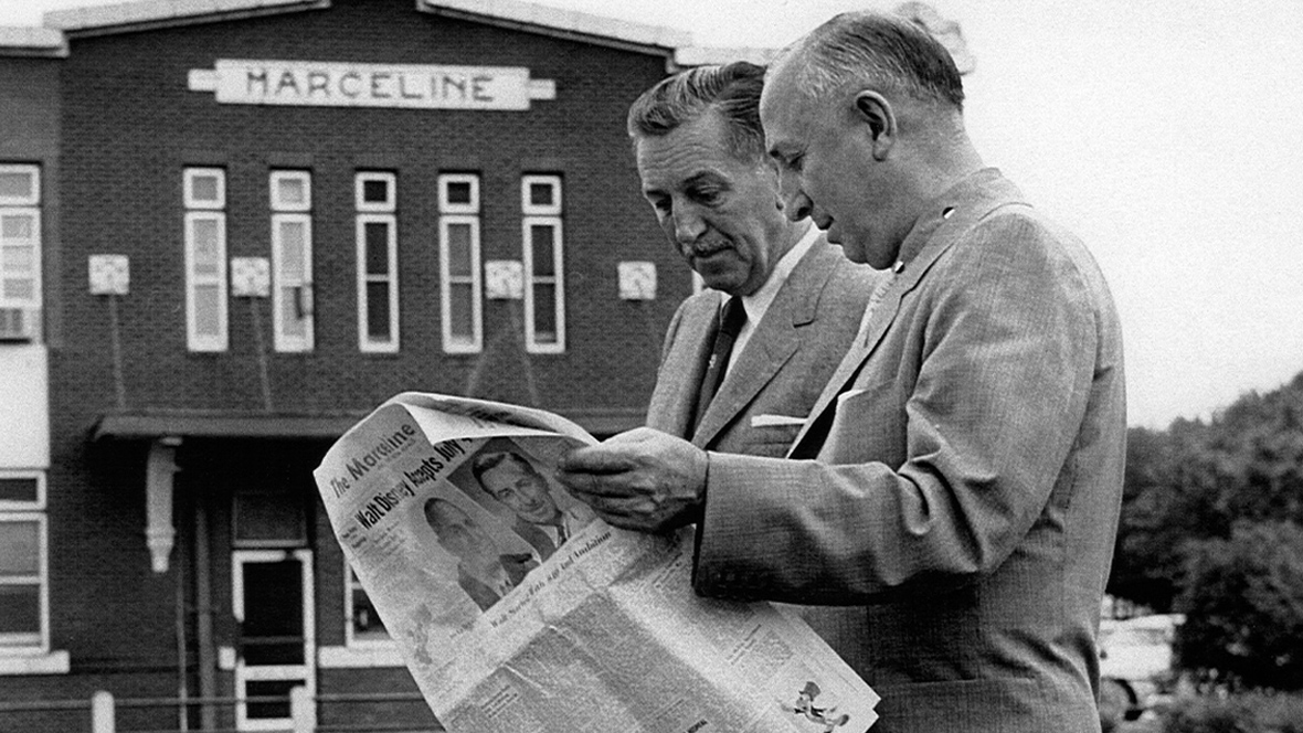 Walt Disney and his brother Roy O. Disney stand outside the train depot in Marceline, Missouri, reading a local newspaper. The depot behind them displays the name “Marceline,” the town where Walt spent part of his childhood and which later inspired Main Street, U.S.A. in Disneyland.