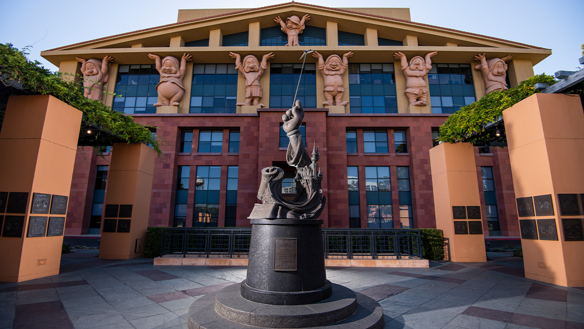 This is the iconic Team Disney Building at the Walt Disney Studios in Burbank, California. The building is famously known for its facade featuring the Seven Dwarfs from Snow White and the Seven Dwarfs holding up the roof, representing their foundational role in Disney’s success. The sculpture in the foreground is “The Legends Plaza” statue, symbolizing the magic of Disney's storytelling legacy