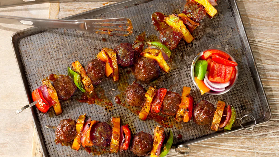 Colorful Hawaiian BBQ skewers featuring golden-brown Impossible® Meatballs, charred pineapple chunks, red bell peppers, and onions, all glazed with a glossy barbecue sauce and served on a wooden board.