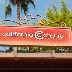 Signage atop the California Churro location. The “California Churro Company” logo sits in white text framed in an orange background. Curled metal surrounds the frame with the Mickey Mouse logo centered at the top. Behind the sign is greenery and a blue-sky background.