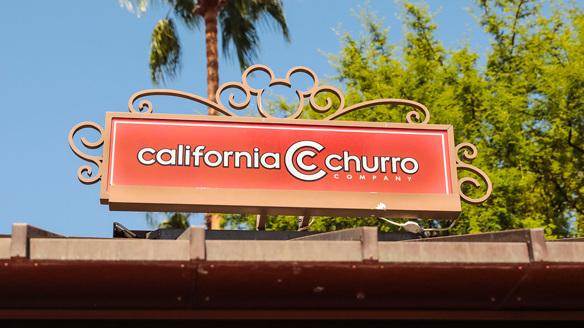 Gold Member Offer: 10% Off at California Churro in Downtown Disney, CA Signage atop the California Churro location. The “California Churro Company” logo sits in white text framed in an orange background. Curled metal surrounds the frame with the Mickey Mouse logo centered at the top. Behind the sign is greenery and a blue-sky background.