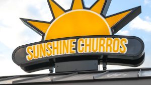 Signage for Sunshine Churros features the company’s name and logo atop the store location on a beautiful sunny day. The yellow Sunshine Churros type is in a yellow font against an oblong shaped black background. Atop the black background is the top half of a yellow sun, framing the logo.