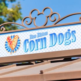 The signage atop of Blue Ribbon Corn Dogs features the company’s logo. Written in blue are the words “Blue Ribbon Corn Dogs” and to the left is an image of two corndogs crossing into an “X”. One corndog is dressed with ketchup and the other with mustard. The signage is housed in a bronze frame with the Mickey Mouse logo topping it.