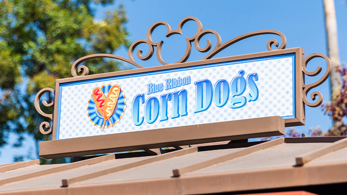 Gold Member Offer: 10% Off at Blue Ribbon Corn Dogs in Downtown Disney, CA The signage atop of Blue Ribbon Corn Dogs features the company’s logo. Written in blue are the words “Blue Ribbon Corn Dogs” and to the left is an image of two corndogs crossing into an “X”. One corndog is dressed with ketchup and the other with mustard. The signage is housed in a bronze frame with the Mickey Mouse logo topping it.