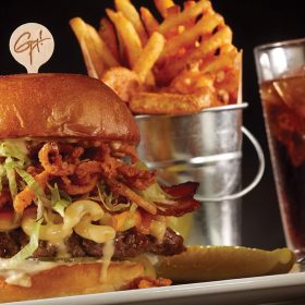 A burger with many toppings, a small metal pail full of fries, and a glass of soda from Planet Hollywood.