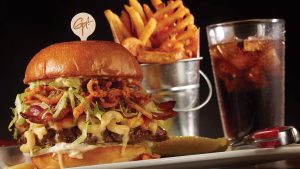 A burger with many toppings, a small metal pail full of fries, and a glass of soda from Planet Hollywood.