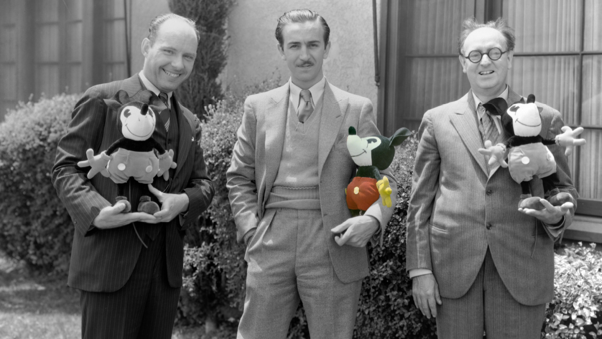 This image shows Walt Disney (center) posing with two colleagues (one on each side), each holding an early Mickey Mouse plush toy. The image is partly colorized to highlight the plush toys' classic red shorts and yellow shoes, while the rest of the photo remains in black and white, giving it a vintage feel.