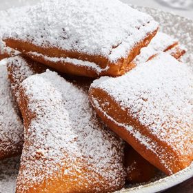 Multiple browned beignets that look as if they’ve just been popped out of the fryer huddle together and rest on a white plate. Mounds of sprinkled white powdered sugar cover each beignet. Some of the leftover powdered sugar sits at the circumference of the plate.