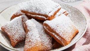 Multiple browned beignets that look as if they’ve just been popped out of the fryer huddle together and rest on a white plate. Mounds of sprinkled white powdered sugar cover each beignet. Some of the leftover powdered sugar sits at the circumference of the plate.