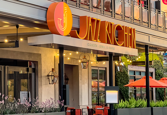 The exterior of the Jazz Kitchen Coastal Grill & Patio in Downtown Disney. The logo sits above the entrance door. Th restaurant has two levels with patio dining on each level. The words “Jazz Kitchen” are written in a thin orange font. To the left of the text is the Jazz Kitchen logo, a circle (half orange, half yellow) with the outline of a saxophone filling the circle. On the patio are a few assorted tables and umbrellas. A stand outside the entrance showcases the menu for those making their dining decision. 
