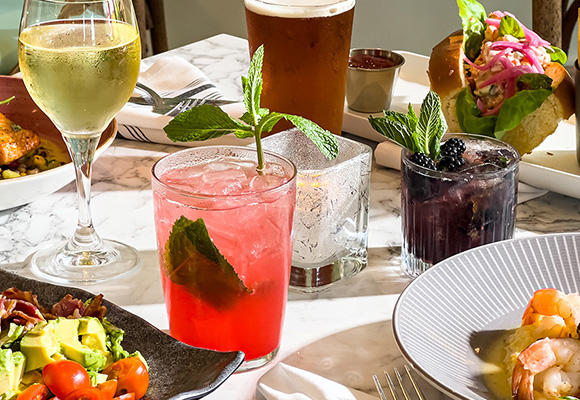 Drinks and food-filled plates sit atop a newly set table. Drinks range from beer to wine to specialty multicolored cocktails, topped with herbs. The food plates include salmon topped with garnish, a salad with bacon, avocado, and tomatoes, a shrimp dish served in a bowl, and a seafood sandwich.