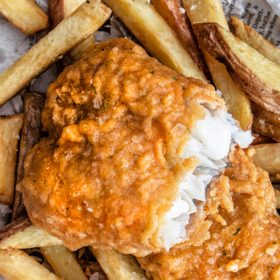 A piece of crispy breaded fish sits, broken in half, surrounded by thick fries and a metal fork. The entire meal sits on wax paper printed to look like a newspaper.