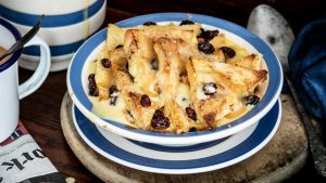 Raglan Road’s Famous Ger's bread and butter pudding sits on a table, housed in a white and blue bowl. The dish is served warm with a hot butterscotch sauce with cool creme anglaise in a cup off to the side.