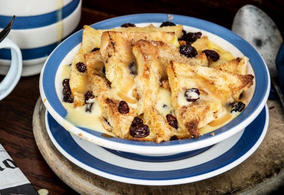 Raglan Road’s Famous Ger's bread and butter pudding sits on a table, housed in a white and blue bowl. The dish is served warm with a hot butterscotch sauce with cool creme anglaise in a cup off to the side. 