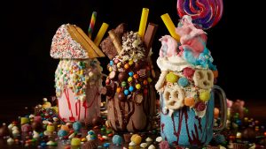 Several Super Nova Shakes from Planet Hollywood are pictured—all three are colorful and topped with piles of candy; the shake on the left has a full piece of cake sitting on top. All three are seen displayed on a large pile of chocolate and candy.