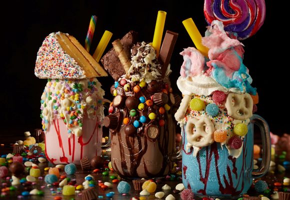 Several Super Nova Shakes from Planet Hollywood are pictured—all three are colorful and topped with piles of candy; the shake on the left has a full piece of cake sitting on top. All three are seen displayed on a large pile of chocolate and candy. 