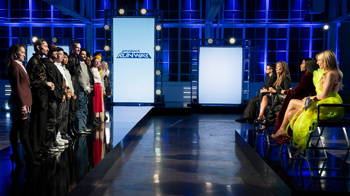 Contestants on Project Runway line up before the judges on a blue-lit runway set, with the show’s logo displayed on tall vertical screens in the background.