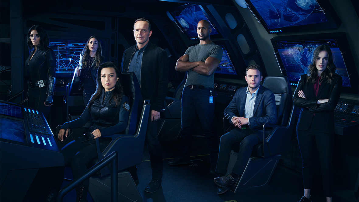 Agents of S.H.I.E.L.D. (2013-20) cast stand in a futuristic blue and black plane. From left to right: Elena Rodriguez, Daisy Johnson, Melinda May, Phil Coulson, Alphonso Mackenzie, Leo Fitz, and Jemma Simmons.