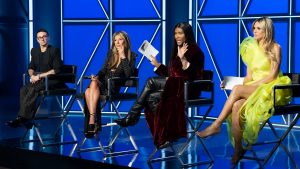 (Left to right) Cristian Siriano, Nina Garcia, Law Roach, and Heidi Klum on the set of Project Runway. They are all sat in black chairs against a blue background. Law Roach is holding up a card and speaking.