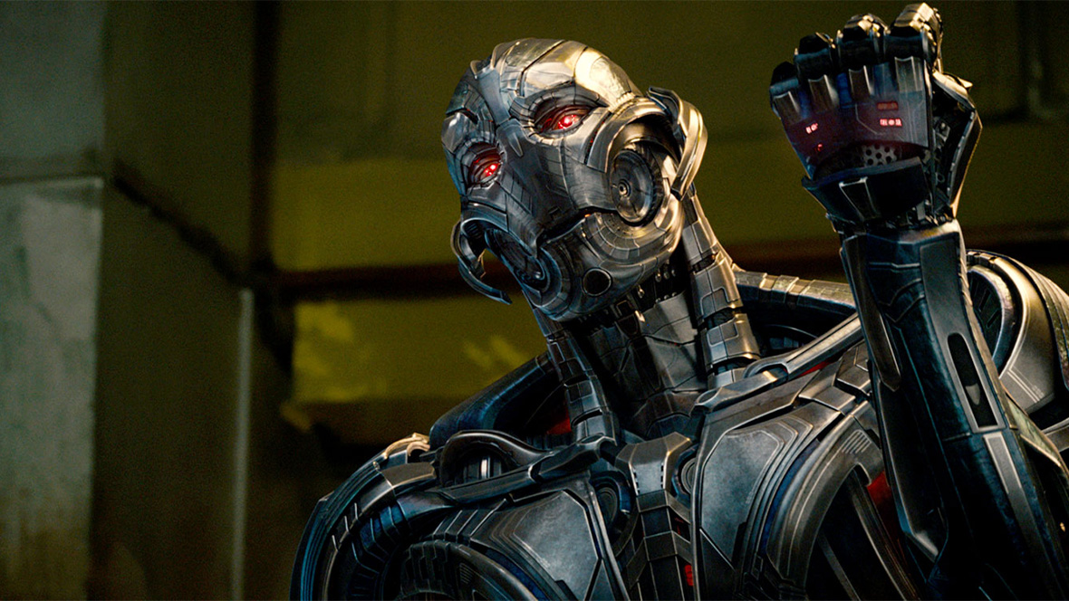 Ultron from Avengers: Age of Ultron (2015) standing in front of a concrete wall and raising a clenched fist.