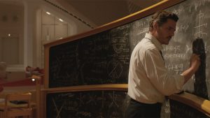 Pedro Pascal as Reed Richards/Mister Fantastic in Marvel Studios’ The Fantastic Four: First Steps. He is wearing a white button-down shirt and doing complex math on a long chalkboard.