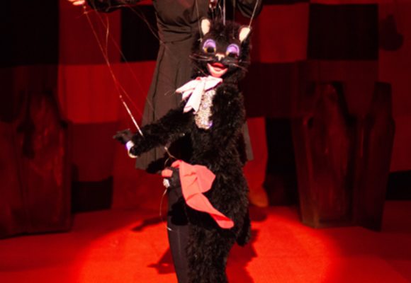 Black Cat marionette dances on red carpet with patchwork curtain behind her. Cat has purple eyeshadow, pink neck scarf, diamond necklace, pink handkerchief, and black high heels. Puppeteer in black dress is visible behind her. 