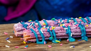 Collage with two images of churros. On the left, a Banana Split Churro rolled in chocolate sugar and topped with banana sauce, vanilla saucer, and a cherry. On the right, a 70th Celebration Churro rolled in ube sugar and topped with marbles blue raspberry icing drizzle and sprinkles.