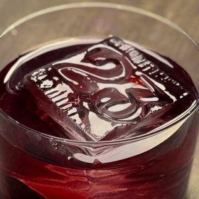 A close up of the No. 23 Cocktail from Frontera Cocina sits in a small cocktail glass on a wooden surface. The specialty drink is a rich dark red color. Floating in the cocktail is a large ice cube that takes up a lot of real estate inside the glass. The square ice cube is stamped with a “23” on it, referring to D23.