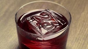 A close up of the No. 23 Cocktail from Frontera Cocina sits in a small cocktail glass on a wooden surface. The specialty drink is a rich dark red color. Floating in the cocktail is a large ice cube that takes up a lot of real estate inside the glass. The square ice cube is stamped with a “23” on it, referring to D23.