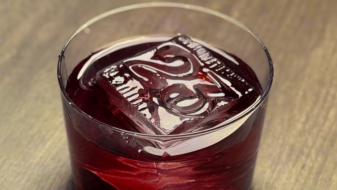 General Member Offer: Special D23 Cocktail at Frontera Cocina in Disney Springs A close up of the No. 23 Cocktail from Frontera Cocina sits in a small cocktail glass on a wooden surface. The specialty drink is a rich dark red color. Floating in the cocktail is a large ice cube that takes up a lot of real estate inside the glass. The square ice cube is stamped with a “23” on it, referring to D23.