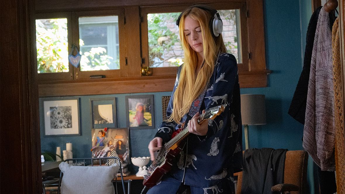 Lindsay Lohan as Anna Coleman in Freakier Friday. She is standing in a home office, wearing headphones and playing the electric guitar.