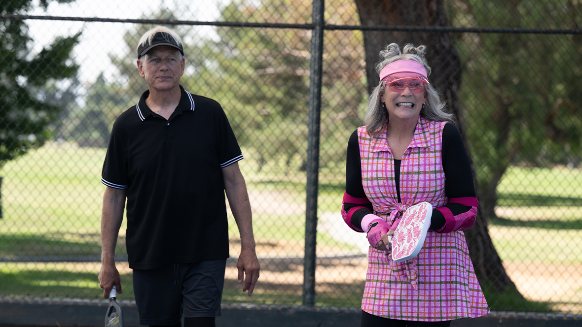 Mark Hammon as Ryan (left) and Jamie Lee Curtis as Tess Coleman (right) in Freakier Friday. The couple is in a park playing pickleball. Ryan is wearing a black visor and black athletic outfit, and Tess is wearing a plaid pink two-piece athletic set with matching elbow pads, sunglasses, and visor.