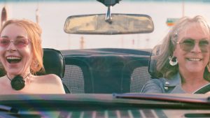 : Lindsay Lohan (left) as Anna Coleman and Jamie Lee Curtis (right) as Tess Coleman in Disney’s Freakier Friday. They are driving a convertible while wearing large colored sunglasses.
