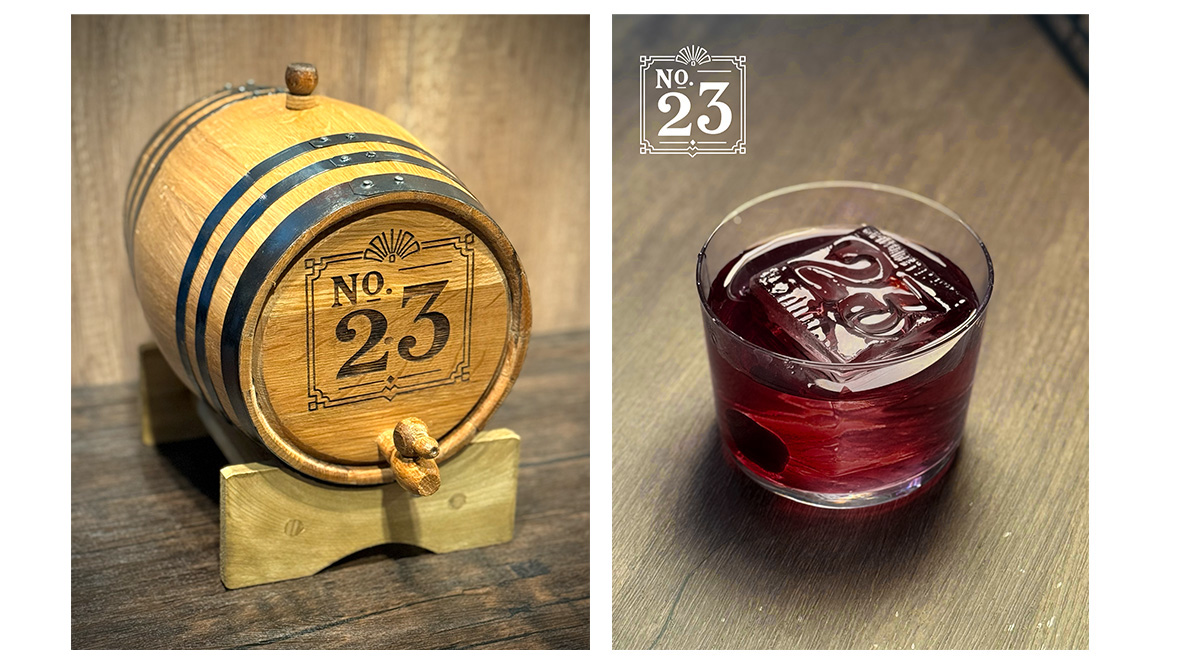 A cocktail glass with red liquid and an ice cube stamped with “No. 23,” next to a wooden barrel labeled the same.