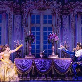 Kyra Belle Johnson as Belle and Fergie L. Philippe as Beast in an exclusive photo from Disney Theatrical Group’s North American Tour of Beauty and the Beast. They are seen in the dining room of the Beast’s castle; Belle is wearing her iconic yellow ball gown. Both are lifting glasses.