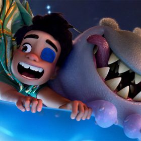 In a still from Disney and Pixar’s Elio, Elio (voiced by Yonas Kibreab), left, and Glordon (voiced by Remy Edgerly), right, are excitedly riding in a light blue cup-shaped vehicle through the Communiverse. They’re holding onto the vehicle for dear life. Elio has a patch over one eye and his mouth is open in a wide smile; Glordon’s tongue is sticking out of one side of his mouth.