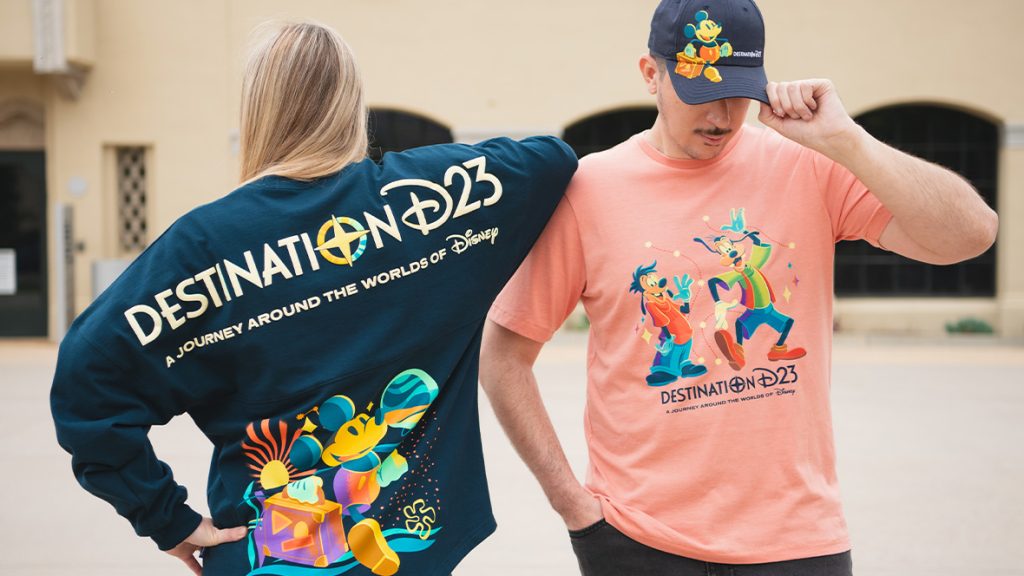 A woman (left) and a man (right) are modeling Destination D23 merchandise; the woman is wearing the Destination D23 spirit jersey, and she’s standing facing the back to better display the logo as seen on the back of the dark blue jersey, which features Mickey Mouse. The man is wearing a salmon-colored T-shirt with the Destination D23 logo and images of Goofy and Max; he’s also wearing a Destination D23 baseball cap. These are items that will be available at The Walt Disney Company Store during the D23 Shopping Spree at Destination D23.