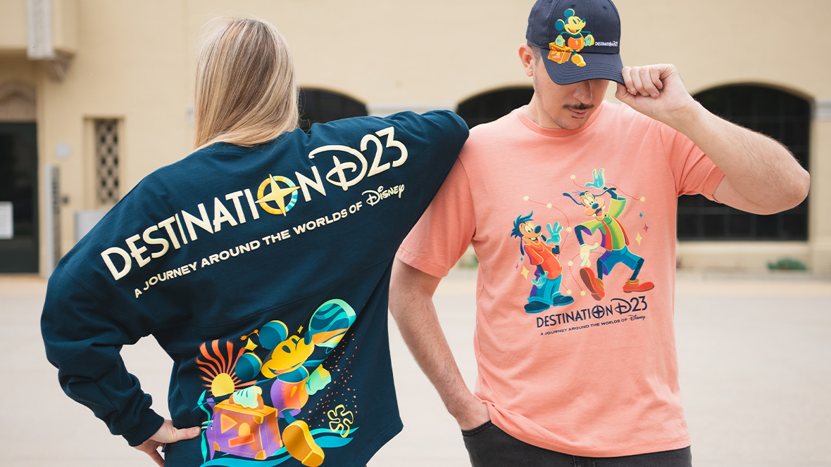 A woman (left) and a man (right) are modeling Destination D23 merchandise; the woman is wearing the Destination D23 spirit jersey, and she’s standing facing the back to better display the logo as seen on the back of the dark blue jersey, which features Mickey Mouse. The man is wearing a salmon-colored T-shirt with the Destination D23 logo and images of Goofy and Max; he’s also wearing a Destination D23 baseball cap. These are items that will be available at The Walt Disney Company Store during the D23 Shopping Spree at Destination D23.