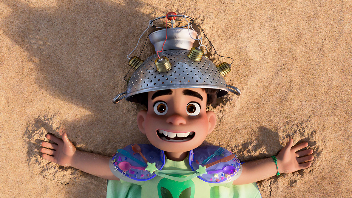 In a still from Disney and Pixar’s Elio, 11-year-old Elio (voiced by Yonas Kibreab) is lying on the beach wearing a homemade cape, a t-shirt featuring a green alien, and a communication device on his head made out of a pasta strainer and other random parts. He is smiling and looking out toward the sky with his arms outstretched. 