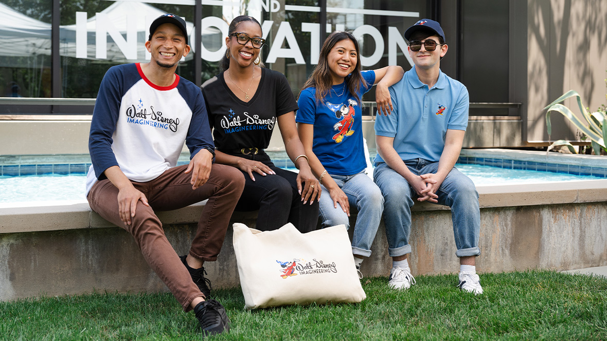 Four people are sitting in front of the small reflecting pool on the Walt Disney Imagineering campus, each wearing and/or displaying WDI merchandise. Among the merch shown are a black WDI logo T-shirt; a WDI logo tote bag; and a blue T-shirt featuring Sorcerer Mickey. These are items that will be available at Mickey’s of Glendale during the D23 Shopping Spree at Destination D23.